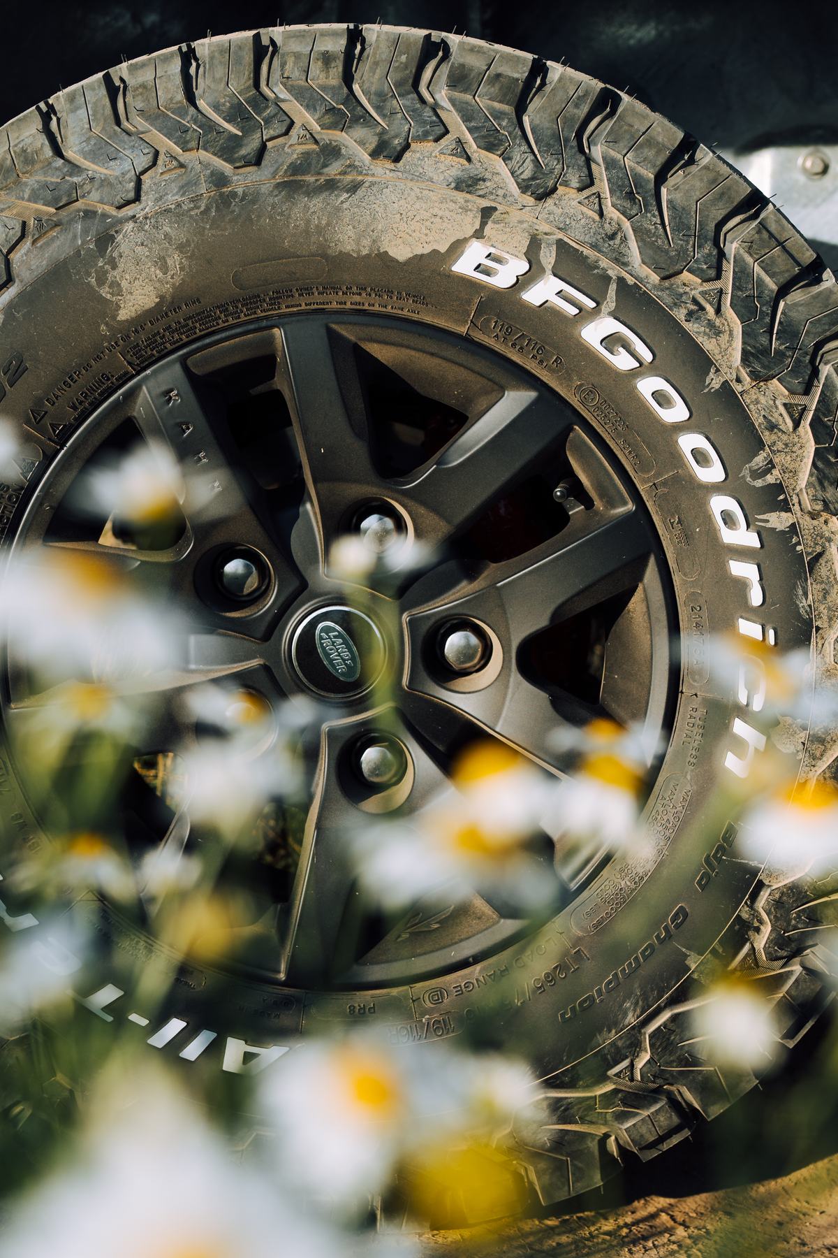 Close-up detail shot of a Land Rover alloy wheel fitted with a muddy BFGoodrich All-Terrain T/A KO2 tyre, photographed through a soft-focus foreground…