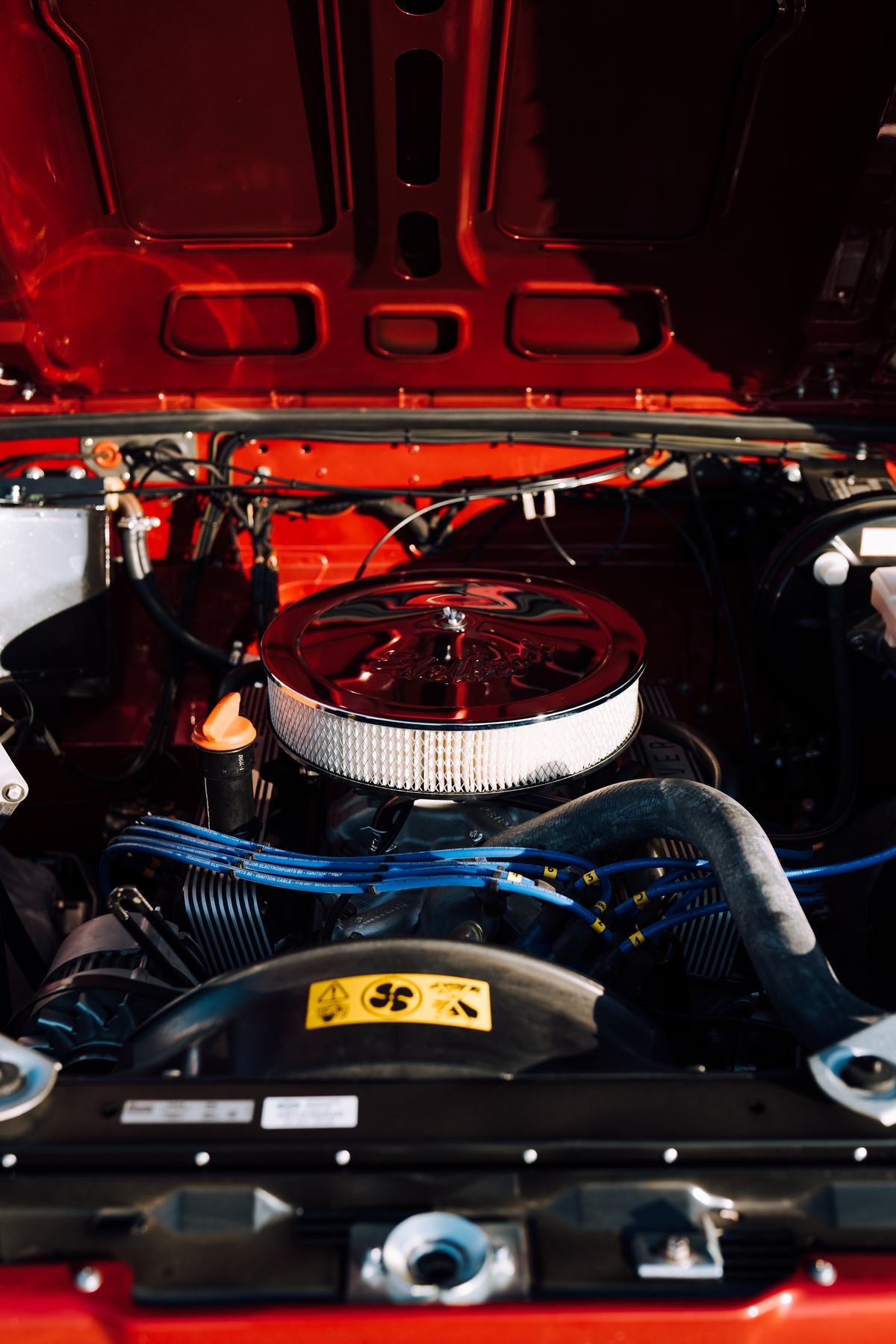 A close-up shot of a classic Land Rover's engine bay with the bonnet open. The vehicle is painted a bright, glossy red. Dominating the centre is a…