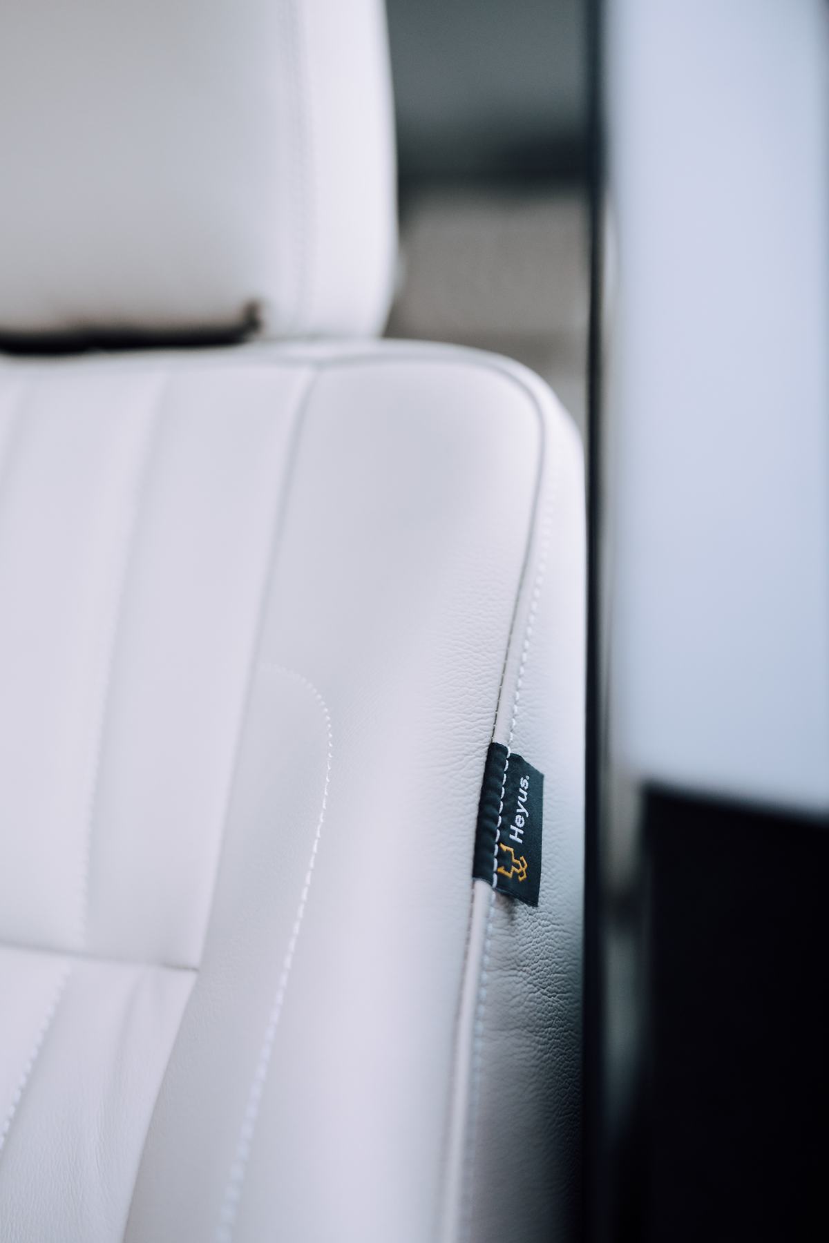 Close-up detail shot of a white leather seat with vertical stitched panels inside a vehicle. A small dark label reading 'Heyus' with a stylised logo i…