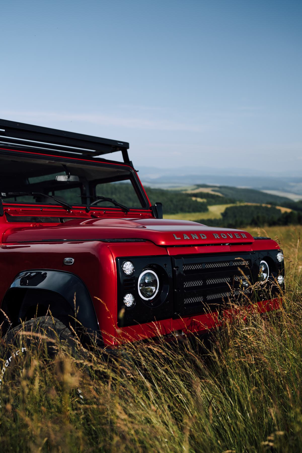A close-up front three-quarter view of a red Land Rover Defender parked in tall grass on a hilltop, with rolling green hills and forest visible in the…