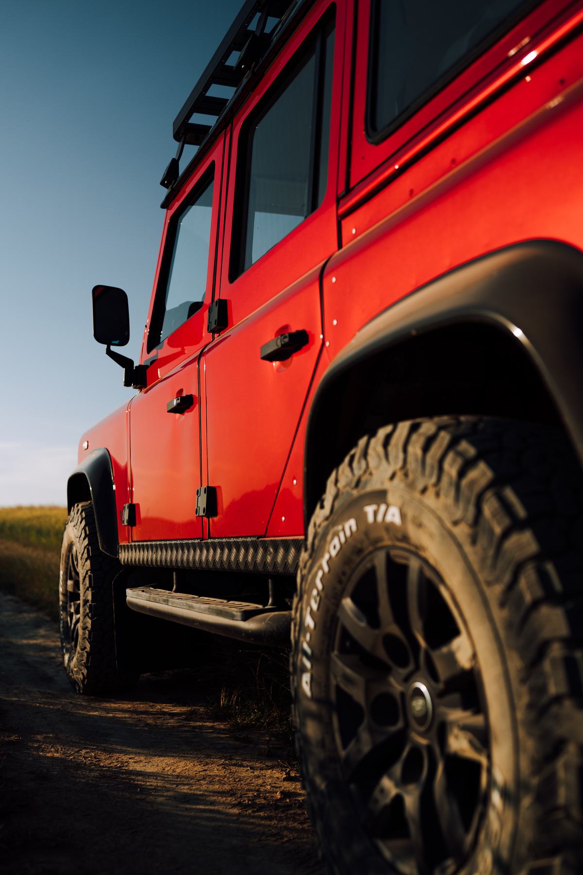 A low-angle shot showing the side of a bright red Land Rover Defender parked on a dirt track. The camera focuses on the side doors and panels,…