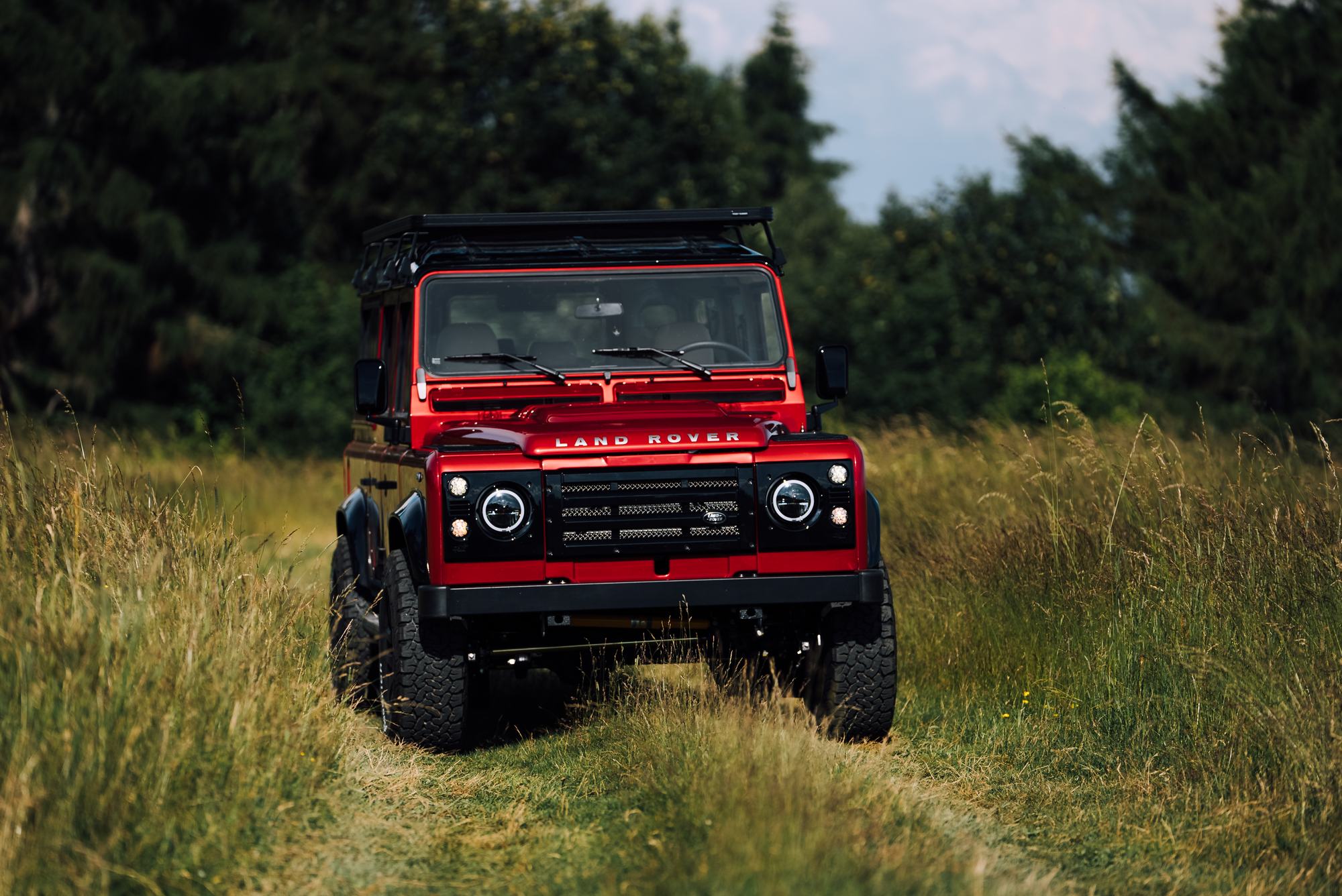 A glossy red Land Rover Defender is parked in a grassy field, viewed from a low front three-quarter angle. The vehicle features a black roof rack,…