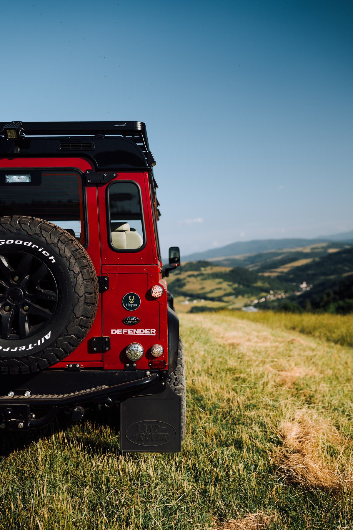 Rear three-quarter detail view of a red Land Rover Defender parked on a grassy hilltop, with rolling hills and a valley visible in the background unde…