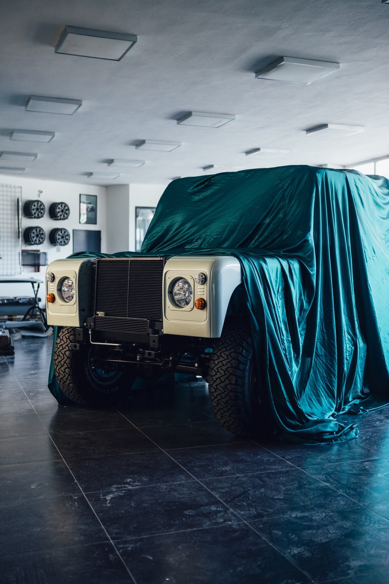 A restored Land Rover with a white front end and exposed grille sits in a showroom, with its body covered by a teal satin sheet leaving only the front…