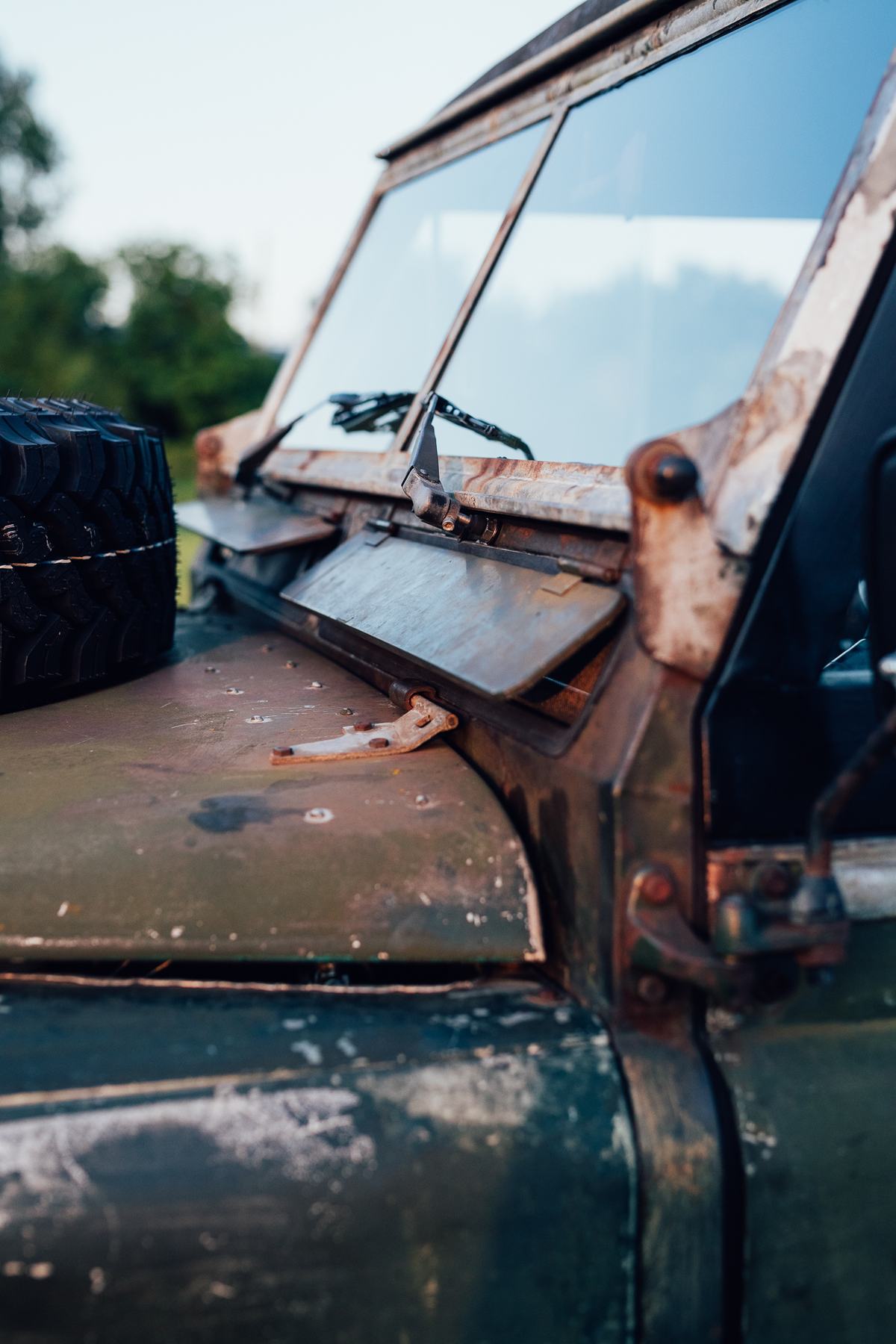 A close-up detail shot of the front bulkhead and windscreen area of a classic Land Rover Series vehicle. The vehicle features deeply weathered,…