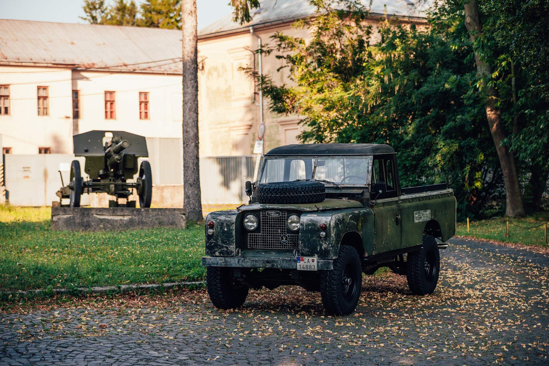 An unrestored, dark green Land Rover Series pickup is parked on a cobblestone surface covered with fallen autumn leaves. In the background, there is…