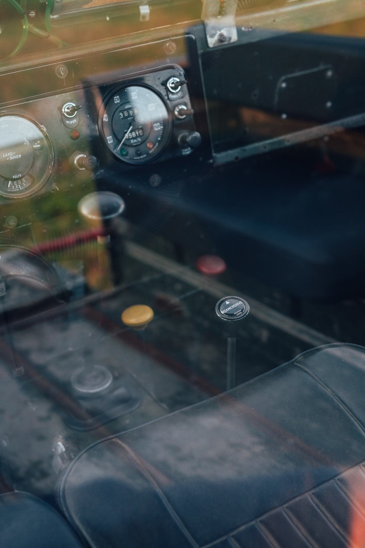 A close-up interior view of a vintage Land Rover Series vehicle, looking through the passenger-side window. The focal point is a black Roamerdrive…