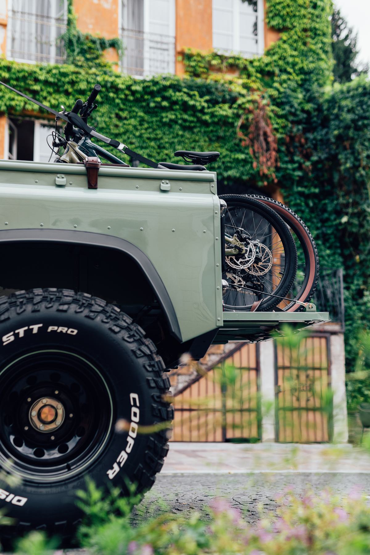 A close-up detail shot of the rear quarter of a finished light green Land Rover Defender pickup, parked on a paved surface outdoors. Two mountain…