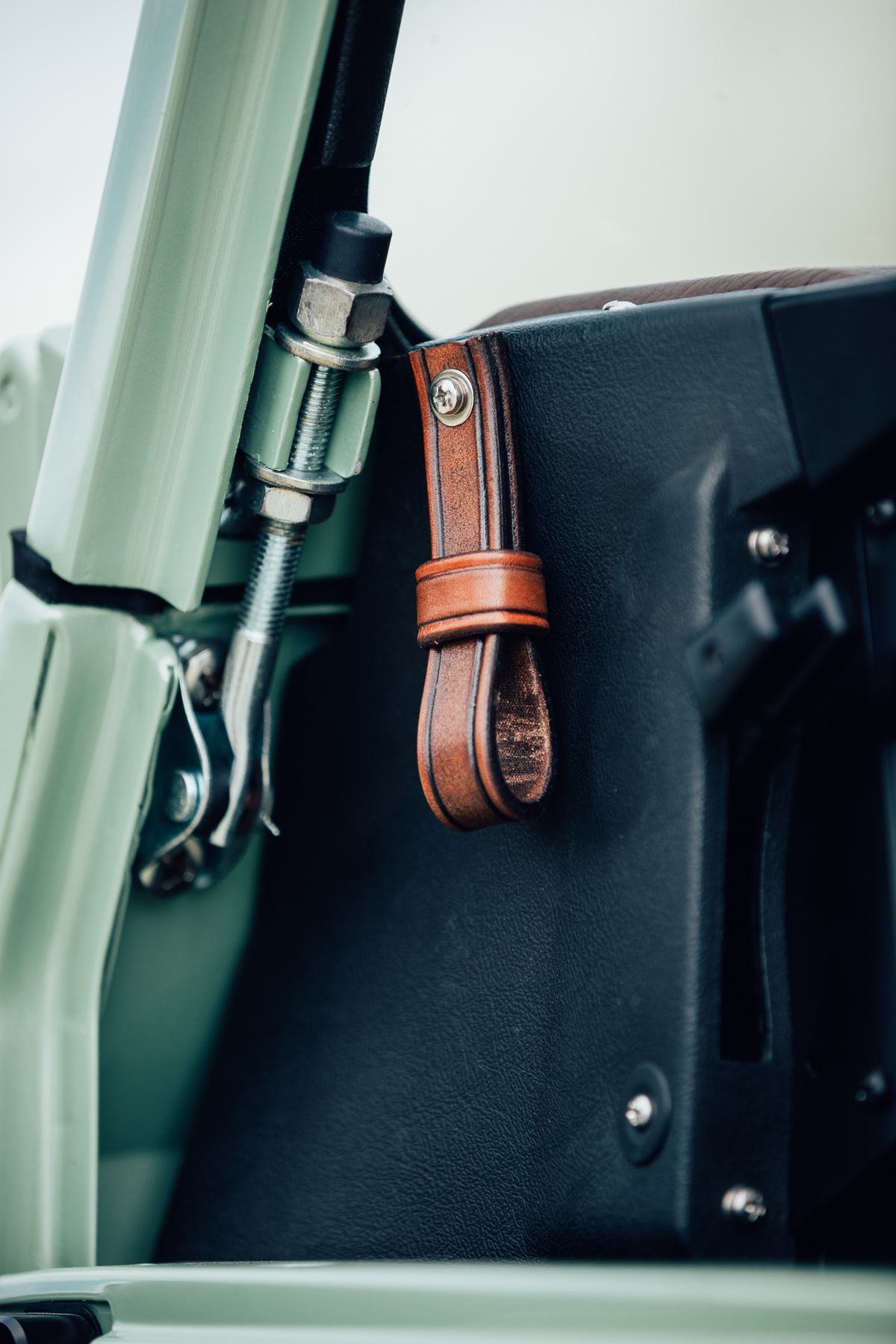 Close-up detail shot of a Land Rover interior, showing a pale green painted A-pillar and windscreen frame alongside a black vinyl or leather-covered s…
