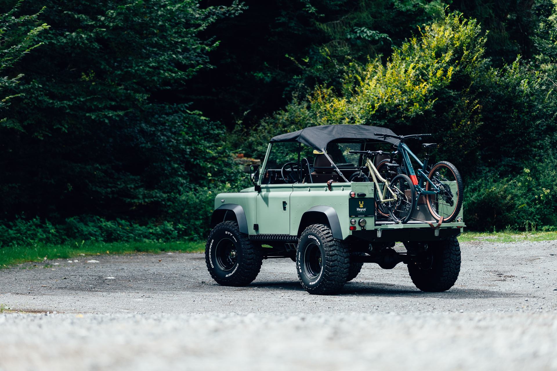 A finished, restored short-wheelbase Land Rover, painted pale sage green with a black soft top and doors removed, is parked on a gravel track in front…