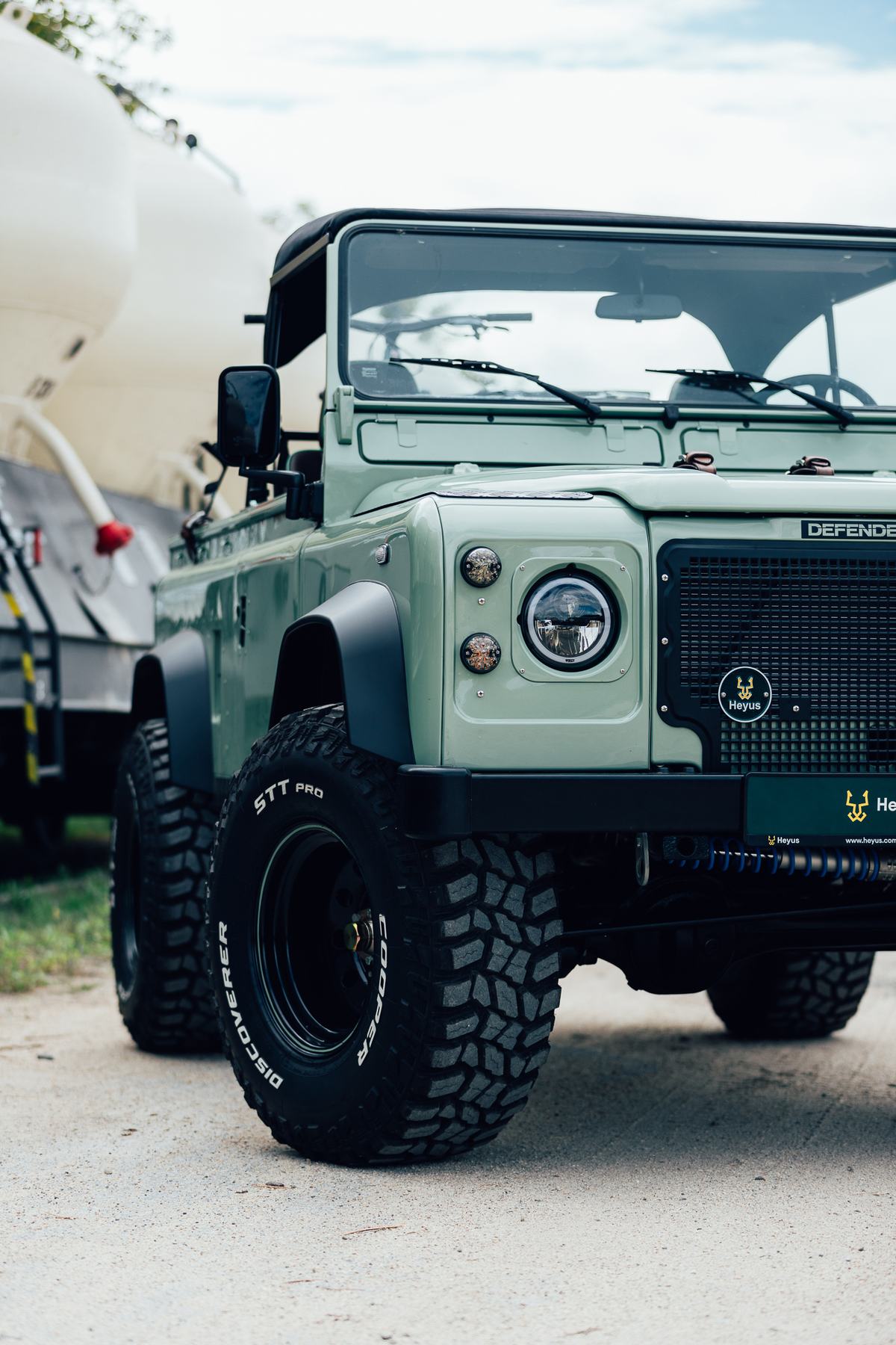 A close, low-angle three-quarter front shot of a restored, soft-top Land Rover Defender finished in pale sage green with matte black wheel arch flares…