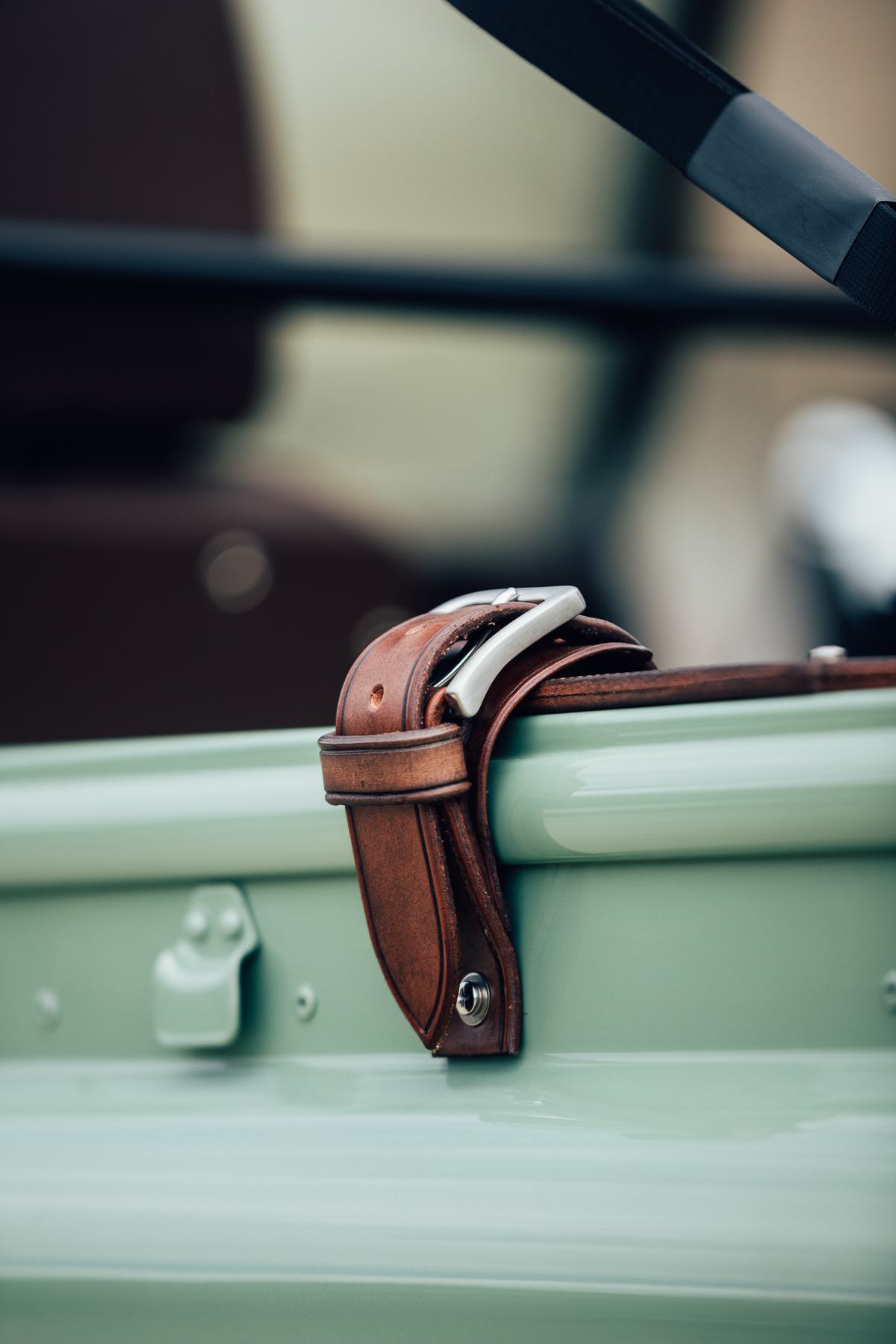 A close-up macro shot of a brown leather strap with a metal buckle, fixed to the edge of a pale green vehicle body panel, likely securing a soft-top o…