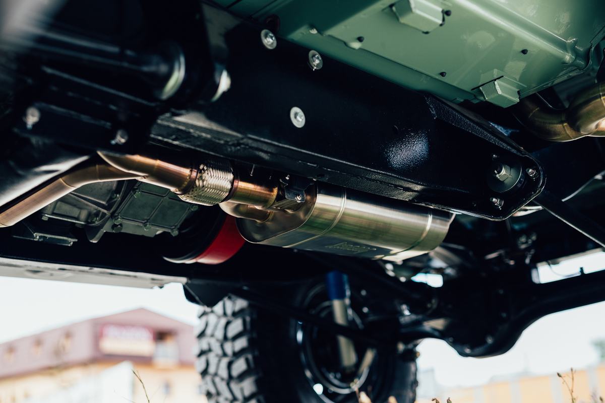 Close-up view from below a Land Rover, showing the underside of the chassis with a stainless steel exhaust system running beneath a black fuel tank gu…