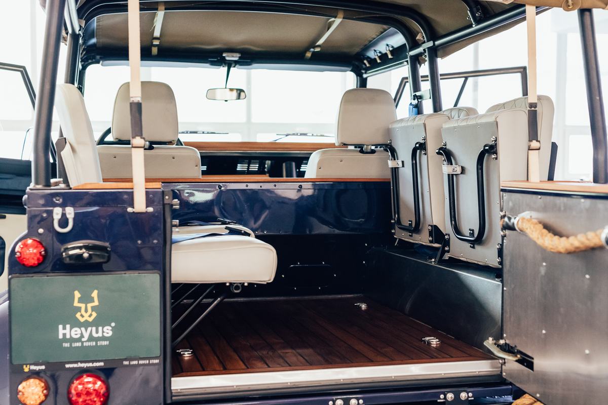 This is an interior shot of a fully restored, blue Land Rover Defender 110 with a soft top, viewed from the open rear tailgate. The vehicle features…