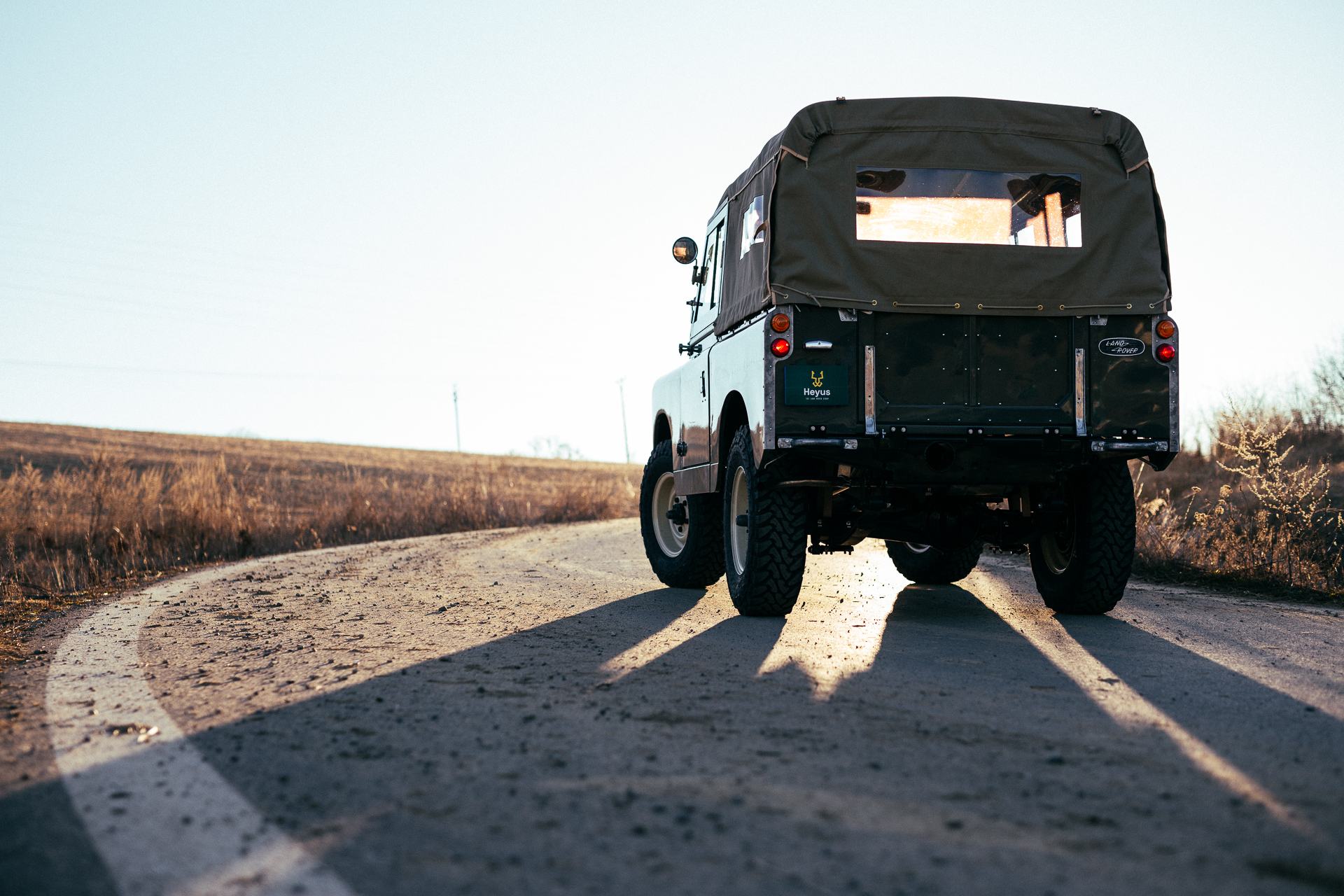 A restored Land Rover Series vehicle with a canvas soft top is photographed from directly behind, parked on a rural tarmac road with dry farm fields o…