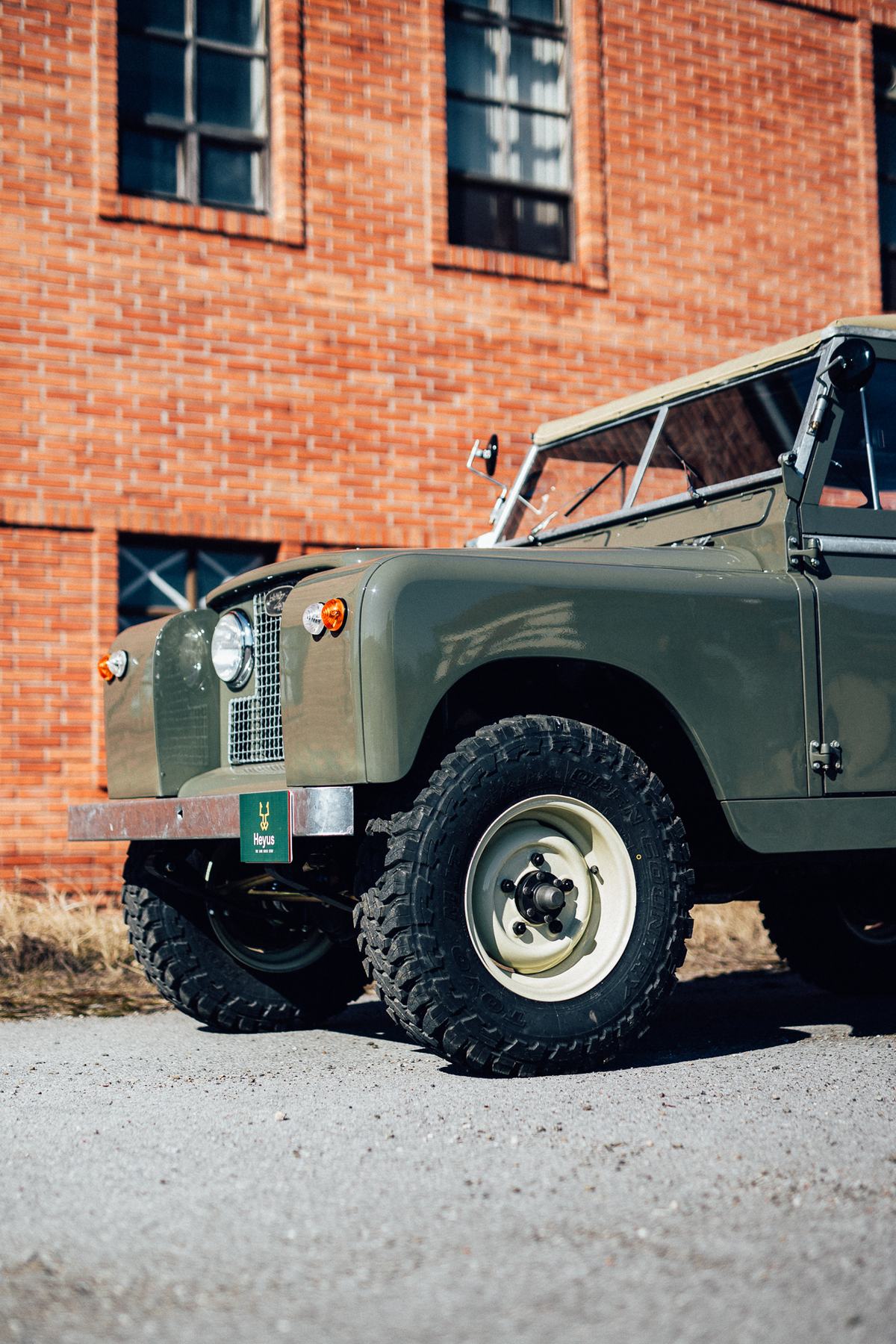 A restored olive-green Land Rover Series with a soft top and canvas roof frame, photographed from a low front three-quarter angle in front of a red br…