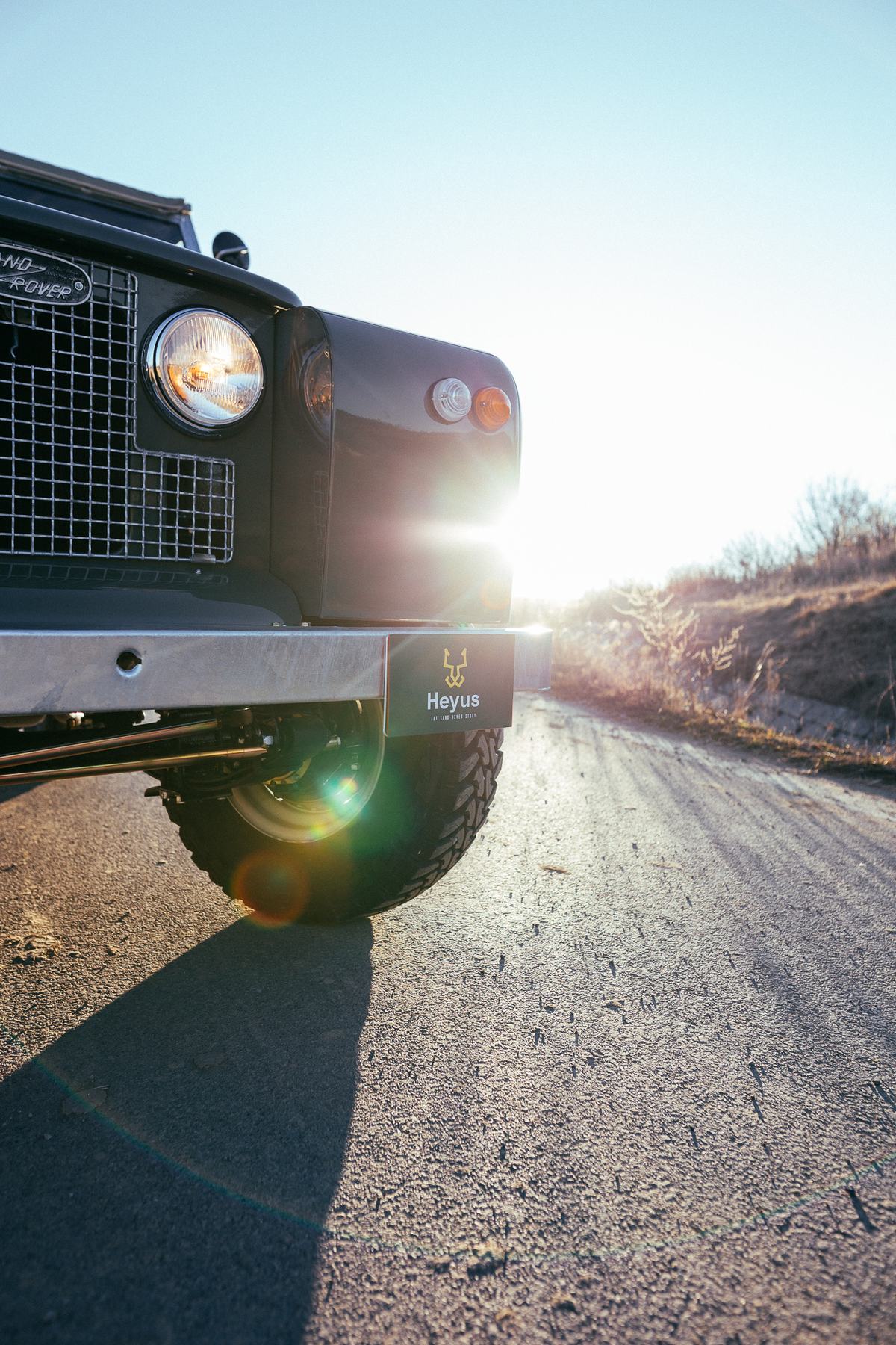 Low, ground-level close-up of the front corner of a dark green Land Rover Series vehicle, shot into low winter sun on a tarmac road. The classic round…