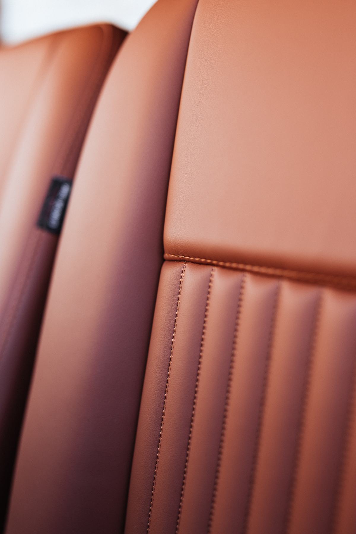 Close-up macro detail of tan/brown leather upholstery, showing seat backs with vertical pleated stitching and a small black fabric label sewn into the…