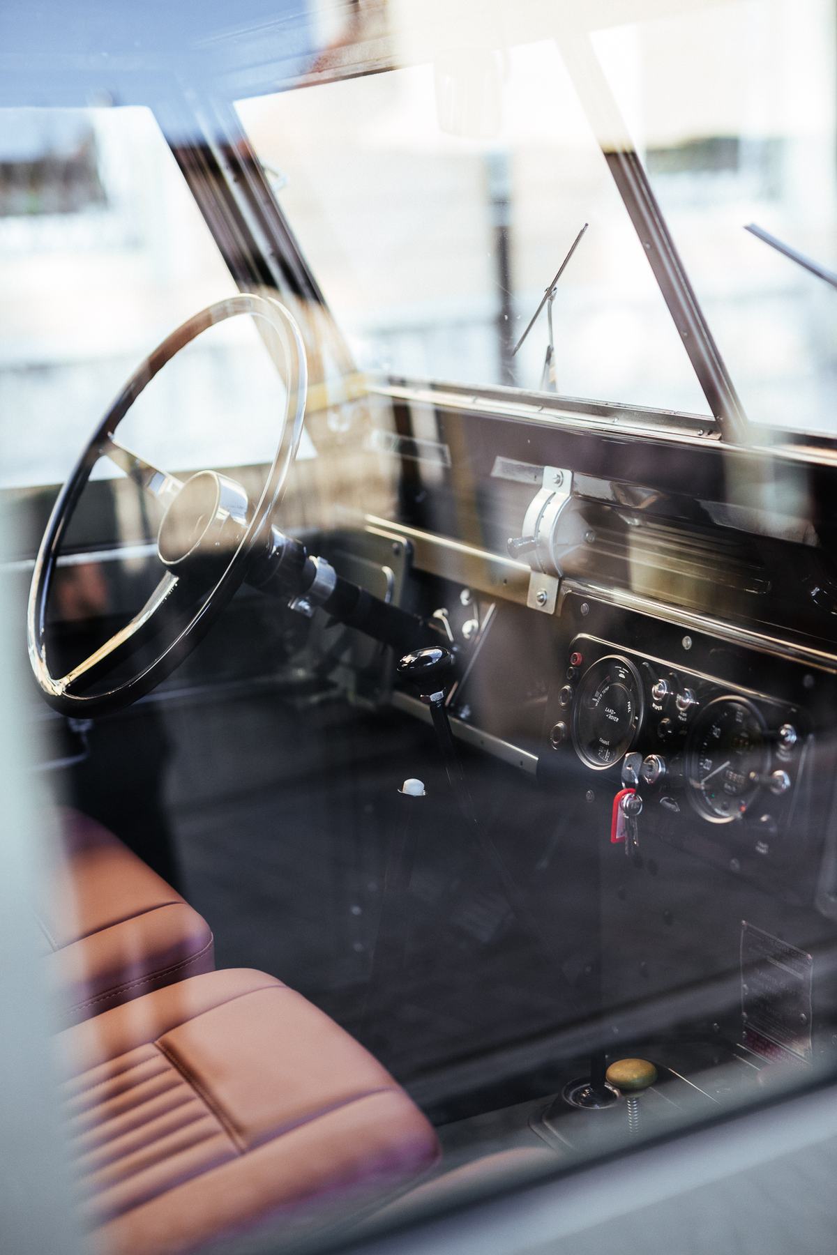 Interior view of a classic Land Rover Series vehicle, photographed through the windshield from outside. A large black steering wheel with a chrome rin…