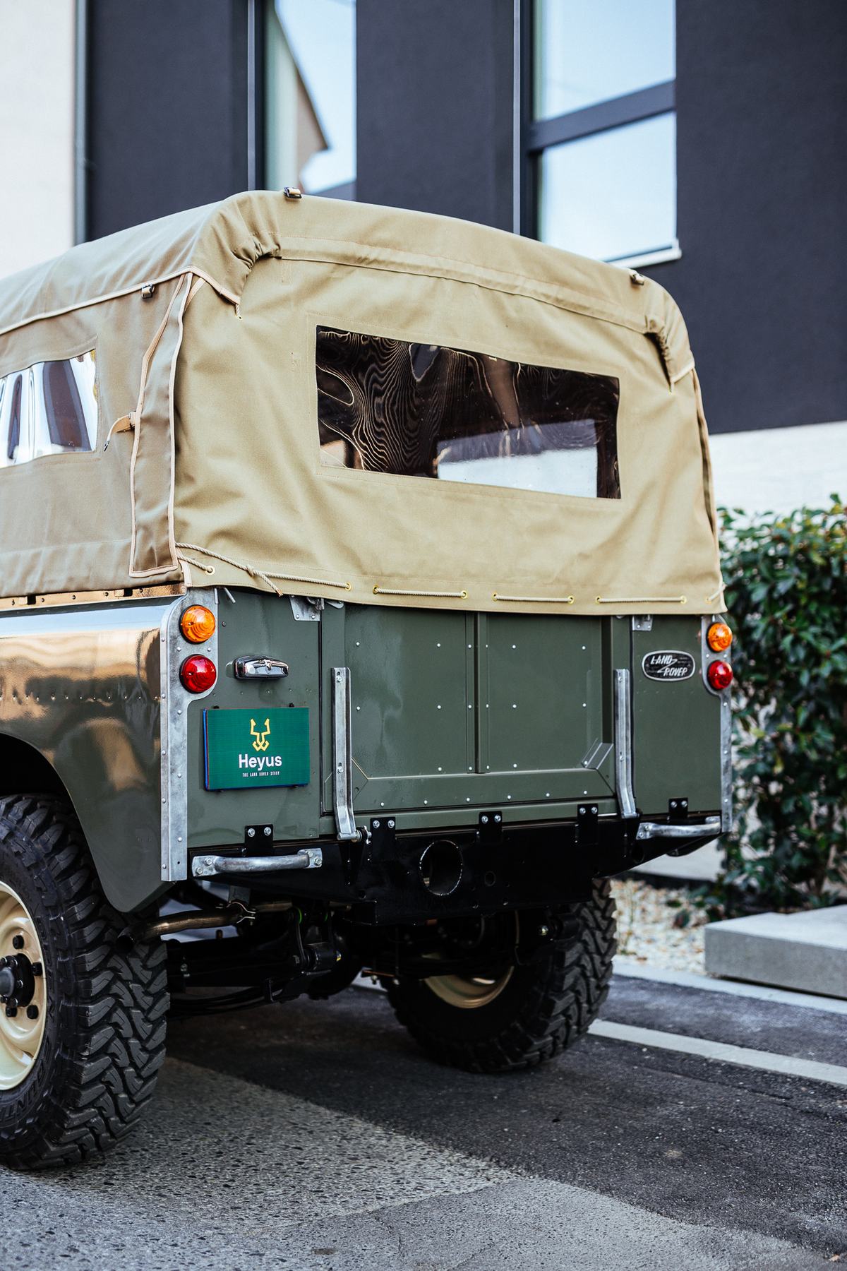 Close rear three-quarter view of a restored green Land Rover Series vehicle with a tan canvas soft top, parked outside a modern building with dark cla…