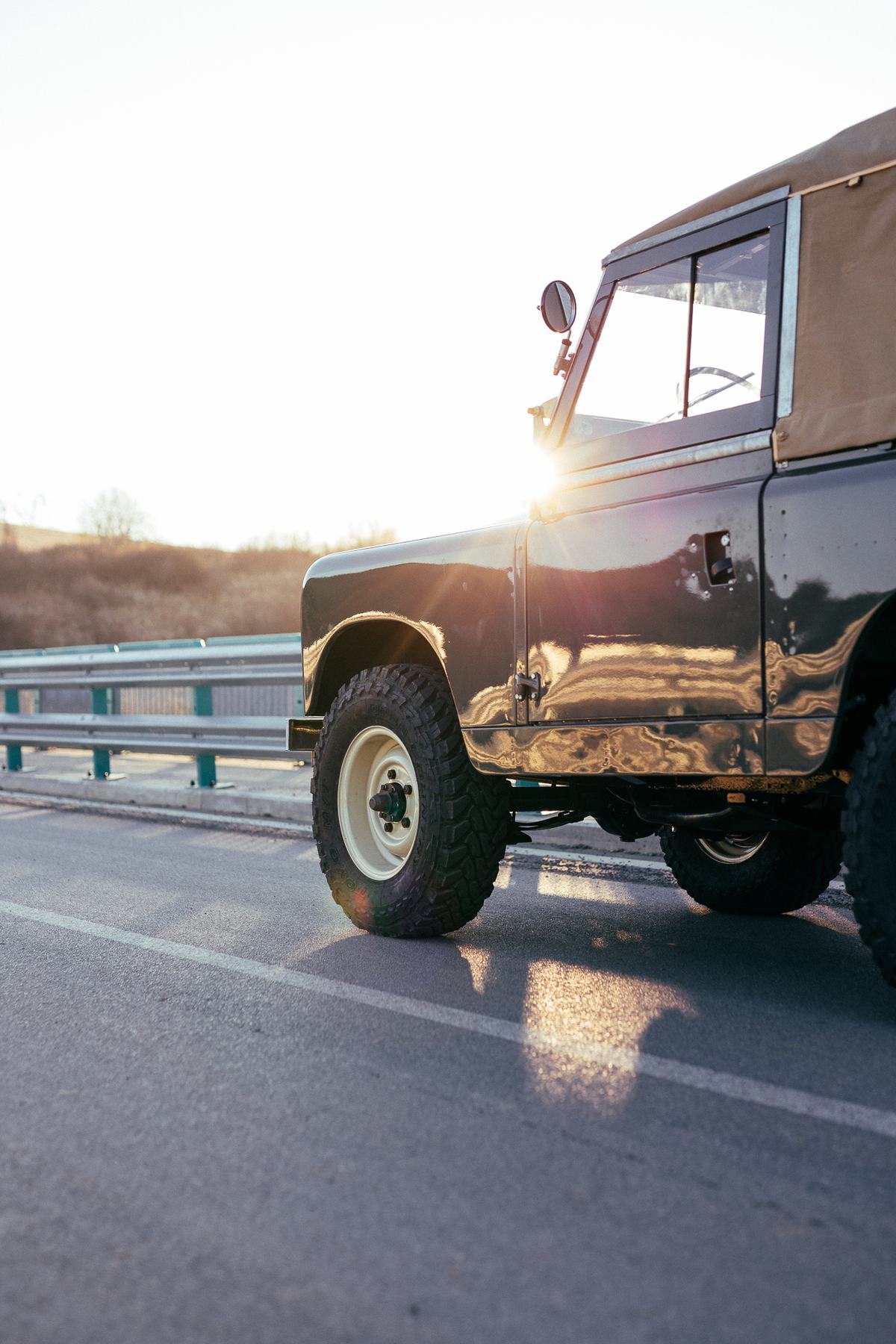 A restored Land Rover Series vehicle with a soft top and glossy dark green paintwork is parked on the shoulder of a road at low sun, with the sun flar…