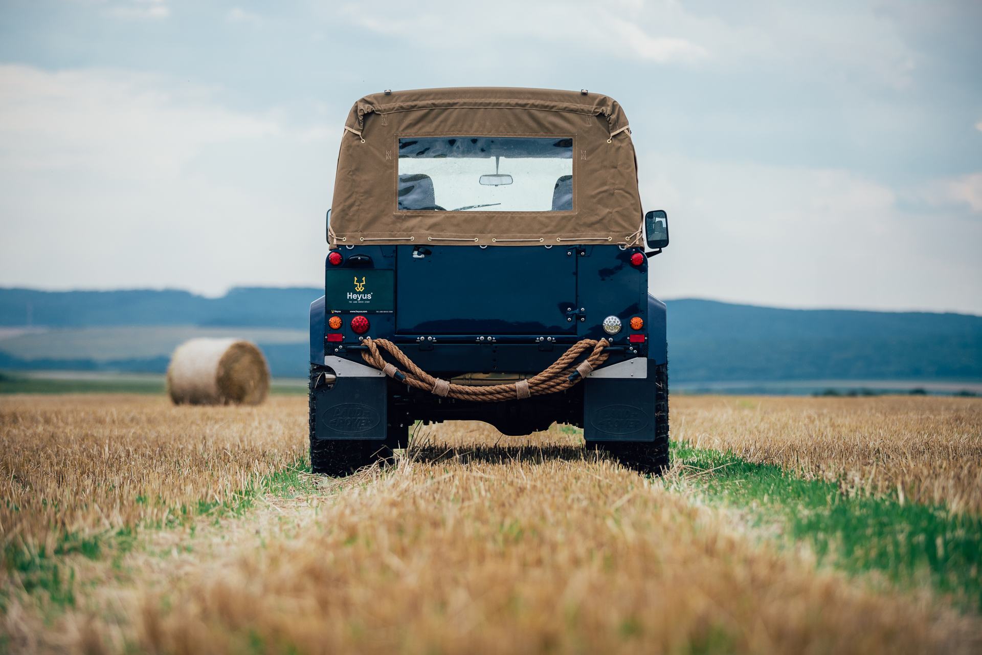 A dark blue Land Rover Series vehicle with a tan canvas soft top is photographed directly from the rear, parked in a harvested wheat field. A thick ro…