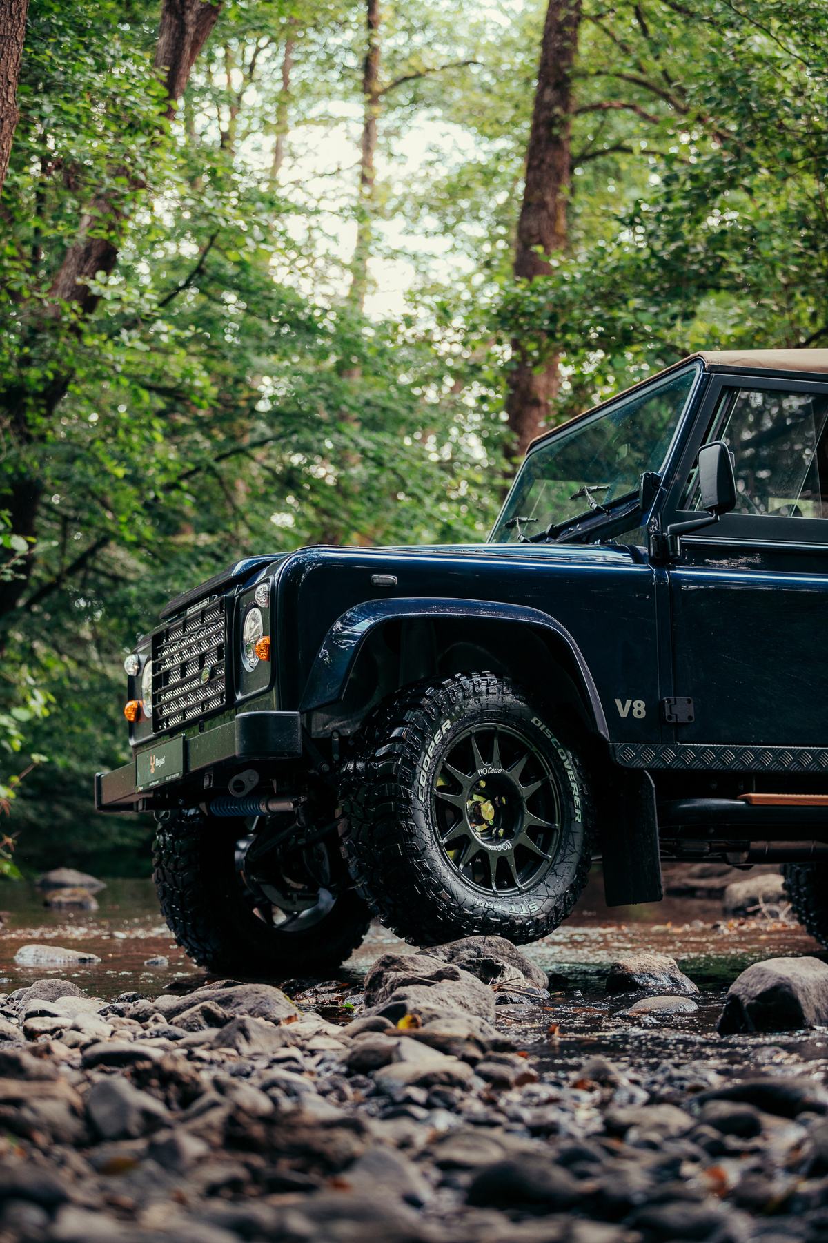 A dark blue Land Rover Defender with a soft top is photographed from a low, ground-level angle beside a rocky forest stream. The truck sits on chunky…