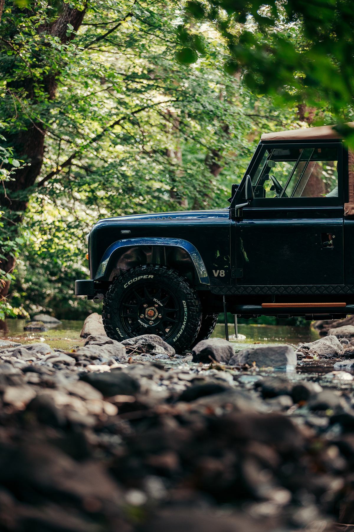A dark blue Land Rover Defender, possibly a 90, is shown partially submerged in a shallow, rocky stream surrounded by a dense forest. The shot is…