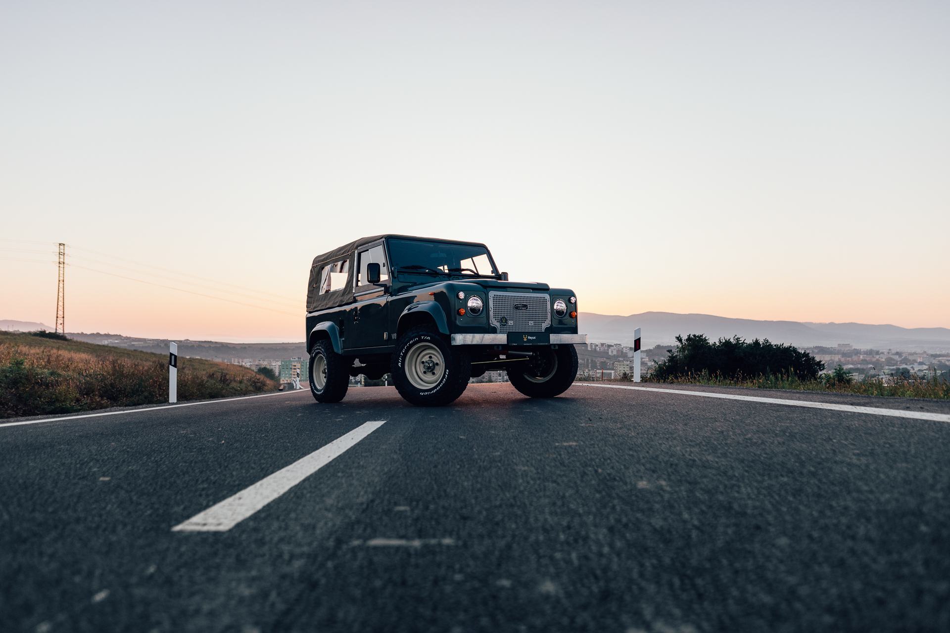 A dark green Land Rover Defender 90 with a soft canvas top is parked on an empty two-lane road at dusk, shot from a low ground-level angle. The vehicl…