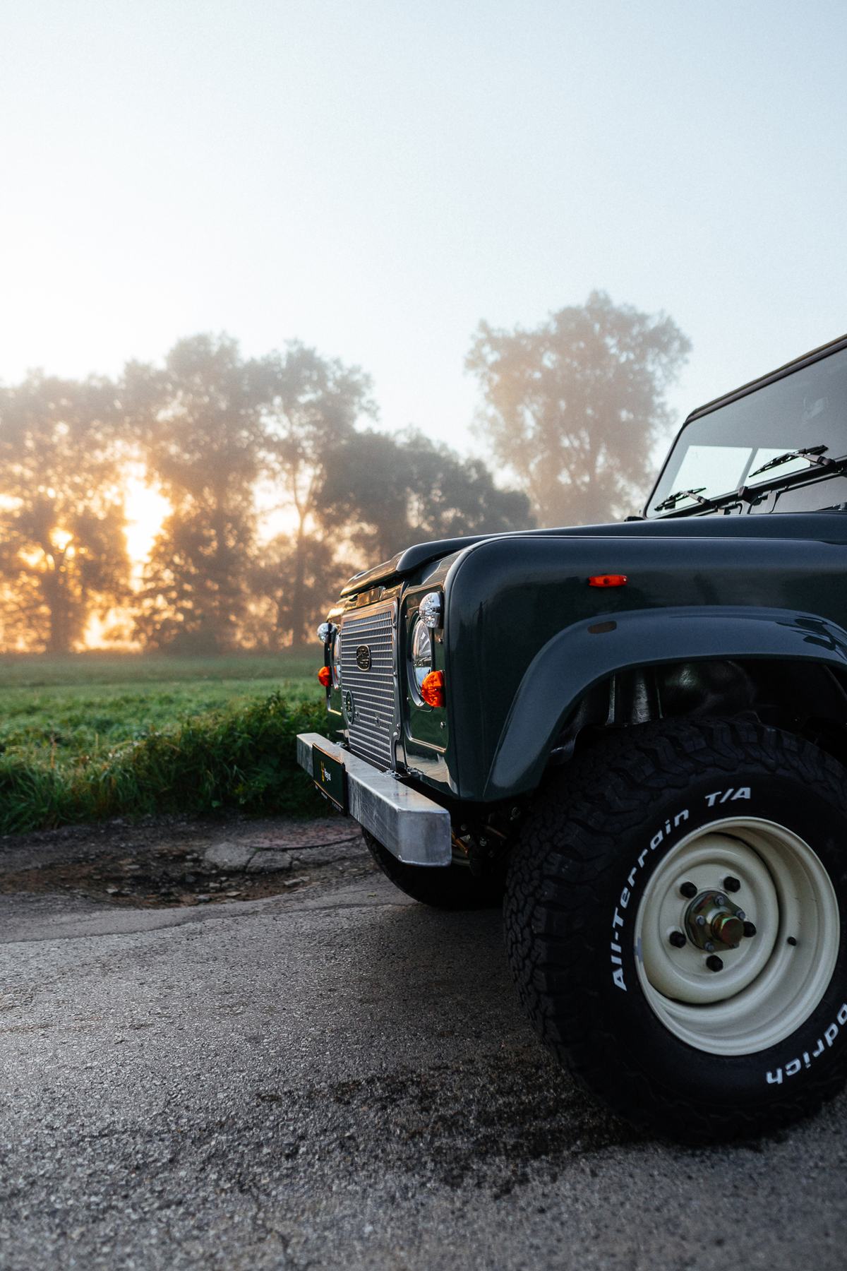 A dark green Land Rover Defender is parked on a gravel track at the edge of a field during a misty sunrise, photographed close-up from the front three…