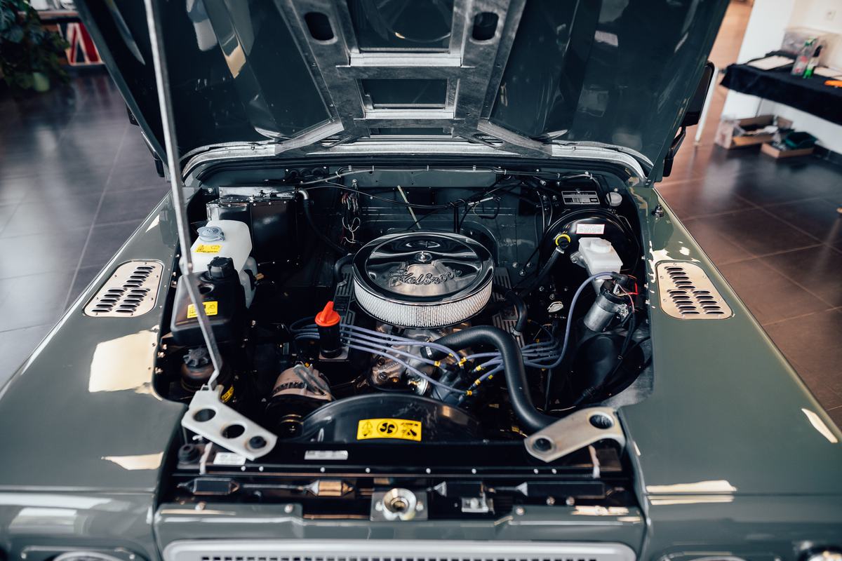 Close-up overhead view into the open engine bay of a dark green Land Rover Defender, photographed indoors in a showroom or workshop. The engine has a…