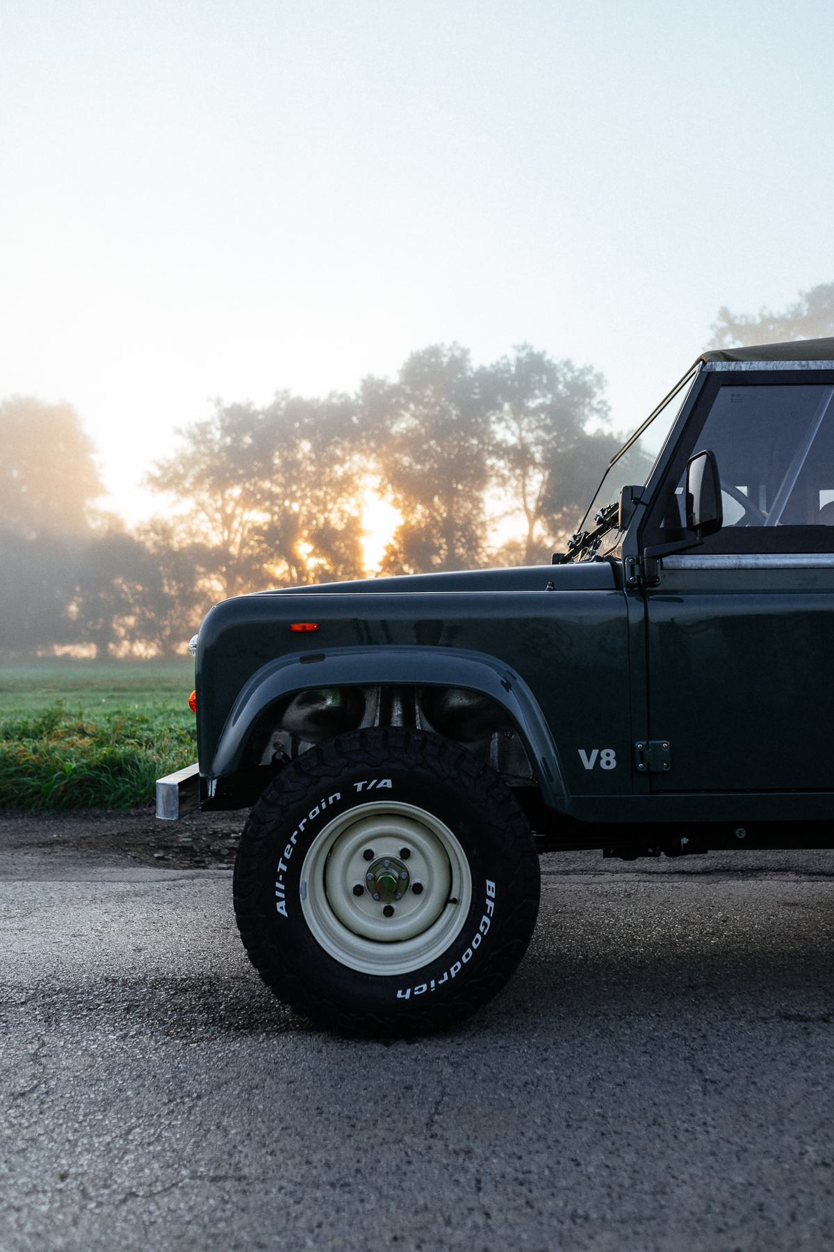 A dark green Land Rover Series vehicle with a soft top is parked on a tarmac road at sunrise, photographed from a low angle focusing on the front whee…