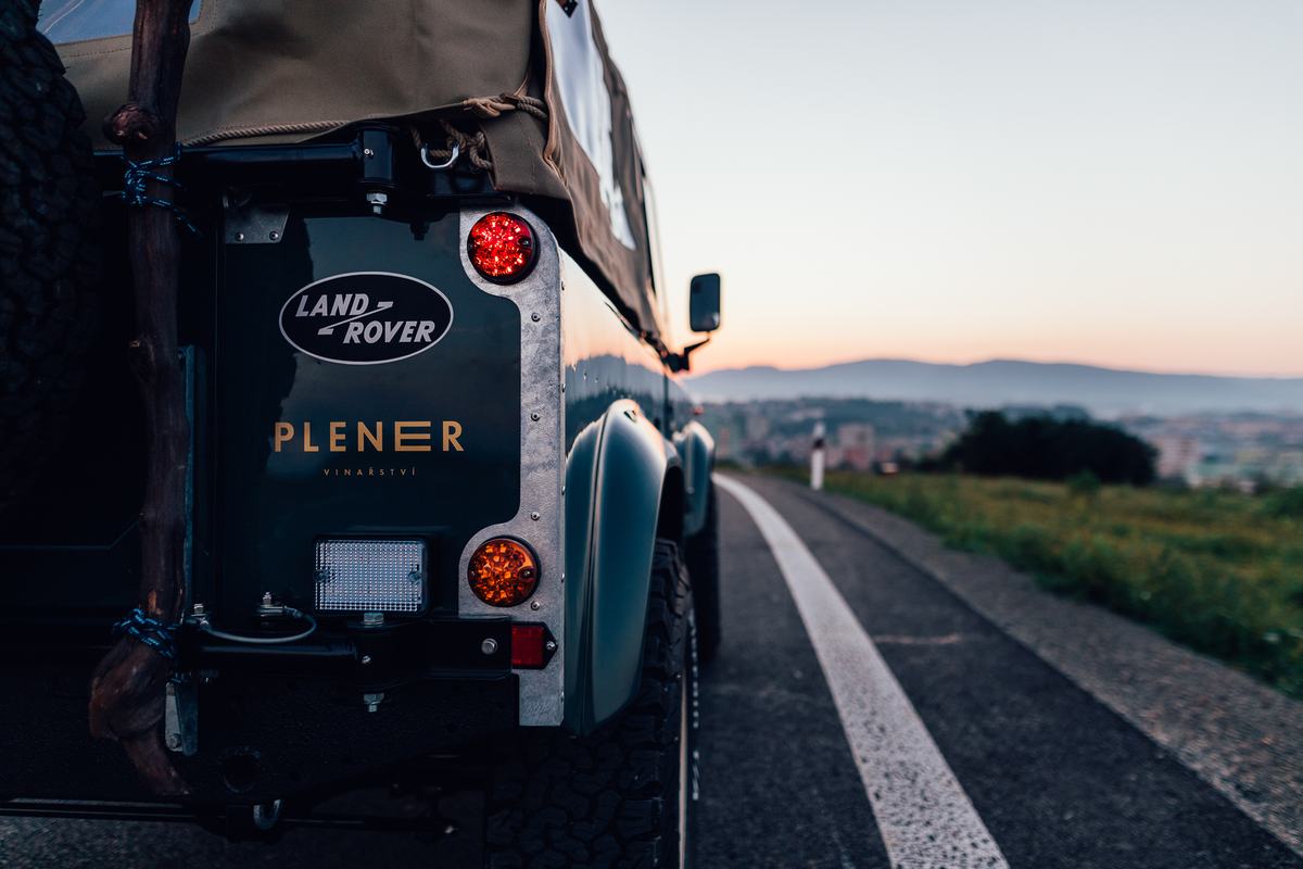 Close-up rear three-quarter detail of a dark green Land Rover Defender parked on a road at dusk, with rolling hills and a distant town visible in the…