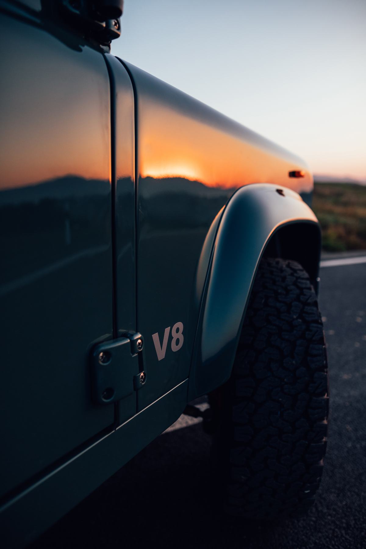 Close-up detail shot of a Land Rover Defender's rear side panel and wheel arch, photographed at sunset. The vivid orange sun and silhouetted hills are…