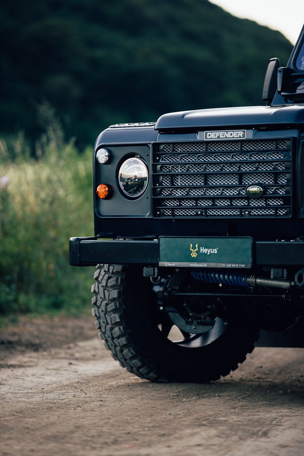 Close-up front three-quarter detail shot of a black Land Rover Defender parked on a dirt track with tall green grass and a dark tree-covered hillside…